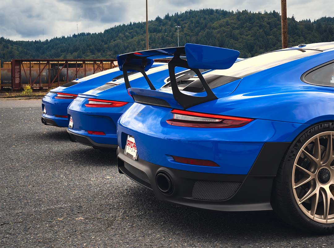 Three Voodoo Blue Porsche 991 GT2 RS sports cars are parked in a row in an outdoor lot, viewed from the rear, with trees and a cloudy sky forming the backdrop.