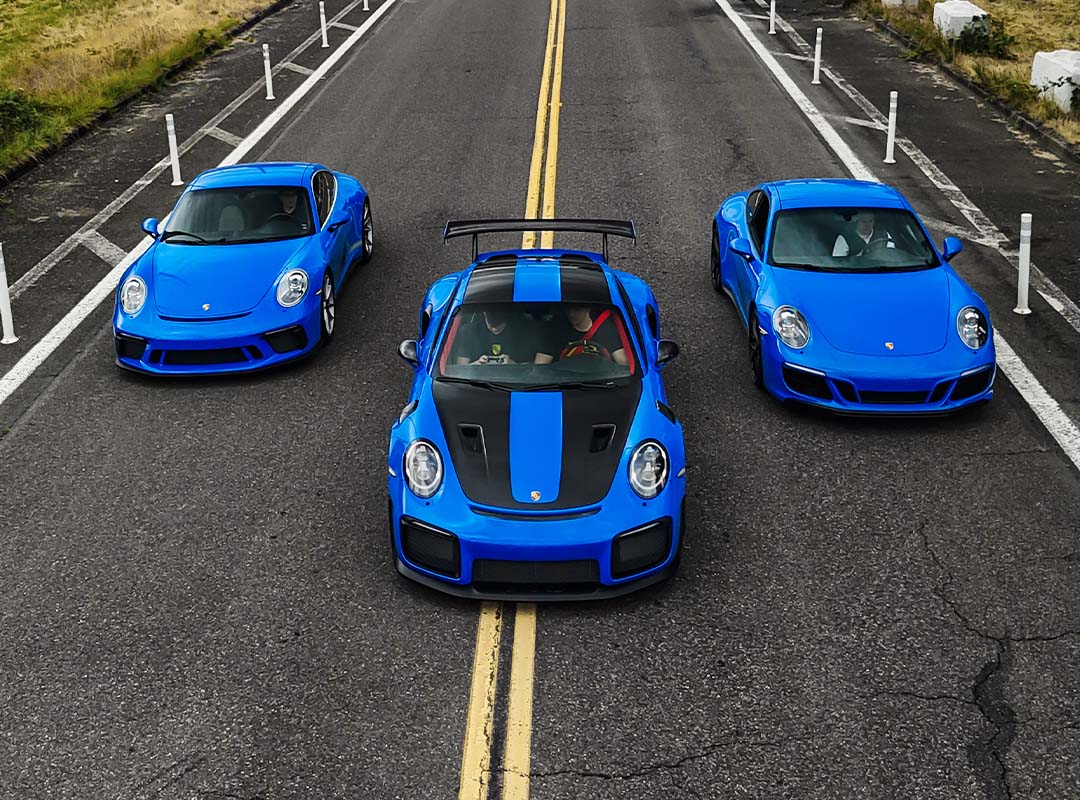 Three blue Porsche sports cars, including a 911 GT3 and a striking GT2 RS, are parked side by side on an empty, two-lane road, viewed from above.