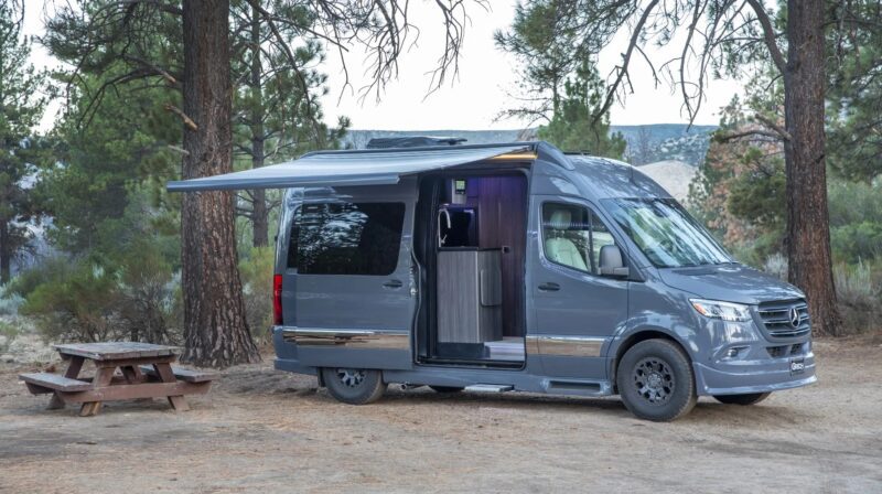 A gray camper van, a unique creation with an extended awning and open side door, is parked at a wooded campsite near a wooden picnic table.