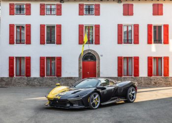 A black and yellow Tailor Made Daytona SP3 Ferrari sports car is parked in front of a white house with red shutters and a red door, capturing the excitement of Car Week Charity Auction.