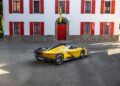 A yellow Ferrari Daytona SP3 sports car is parked in front of a white building with red shutters and a red door, ready to impress at the Car Week Charity Auction.
