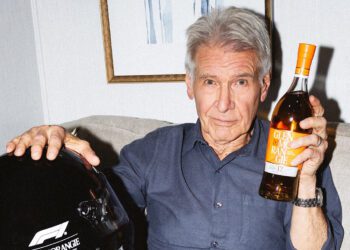 Older man with gray hair sits on a couch, holding a bottle of Glenmorangie whisky in one hand and a sleek Formula 1 racing helmet in the other.