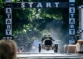 A vintage race car with two drivers begins a race under a "START" banner on a tree-lined track, echoing the excitement of the 10 Fastest Cars At The 2025 Goodwood Festival Of Speed, with hay bales marking the course.