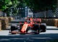 A red Formula 1 car, one of the 10 Fastest Cars At The 2025 Goodwood Festival Of Speed, races on a tree-lined track with hay bales and checkered flags, passing under a "START" banner.