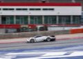 A white Pagani sports car speeds along the Goodwood FoS 2025 race track, with blurred background buildings, barriers, and safety fencing hinting at exhilarating hillclimb runs.
