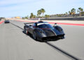 A sleek black Pagani sports car races at high speed on a racetrack lined with palm trees, another car visible in the background during the thrilling 2025 Monterey Car Week.