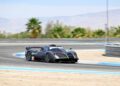 A black Pagani race car with aerodynamic features drives on a curving racetrack during Monterey Car Week 2025, with palm trees and mountains in the background.