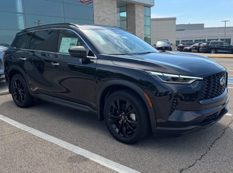 A black Infiniti SUV is parked in a dealership lot on a sunny day, with a building and several cars in the background—perfect for those seeking the best car lease deals.