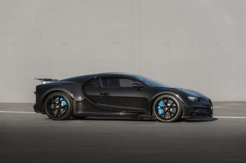 A 2018 Bugatti Chiron black sports car with blue brake calipers is parked on a smooth gray surface, set against a light gray wall—an impressive sight worthy of the Monterey Jet Center Auction.