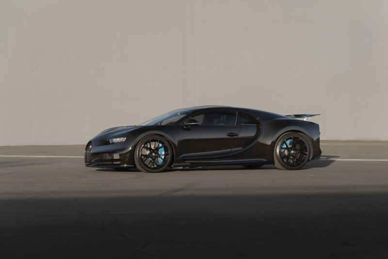 A matte black 2018 Bugatti Chiron sports car is parked on an empty asphalt surface against a plain, light-colored wall.