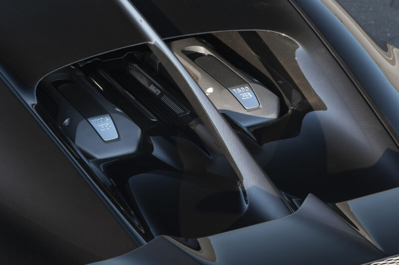Close-up view of a 2018 Bugatti Chiron engine bay at the Monterey Jet Center Auction, featuring two labeled components with metallic covers and sleek black and carbon fiber detailing.
