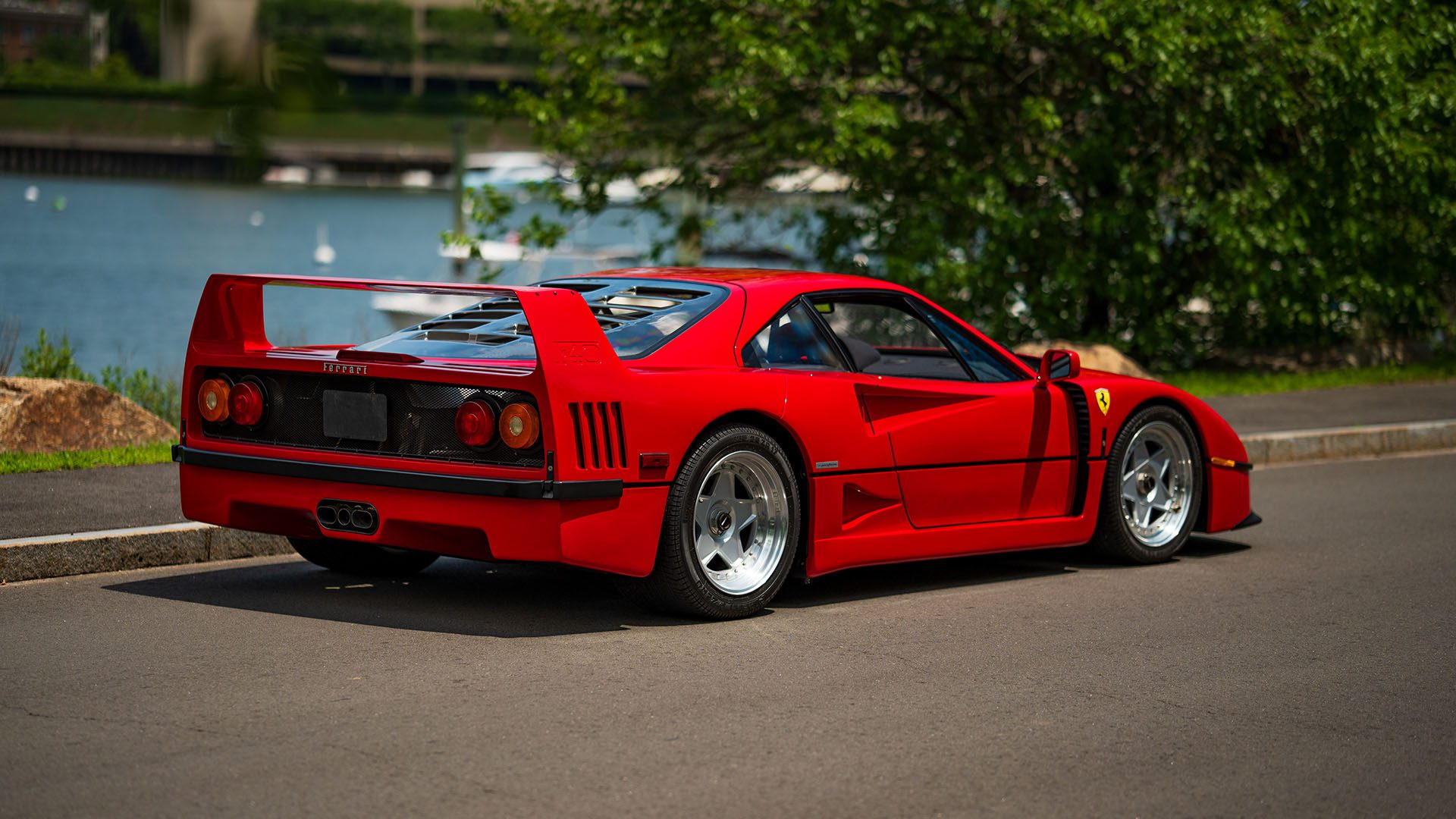 A red Ferrari F40 sports car is parked on a paved road near water and greenery, viewed from the rear left side—an iconic sight reminiscent of the 1991 Ferrari models showcased at the Monterey Jet Center Auction.