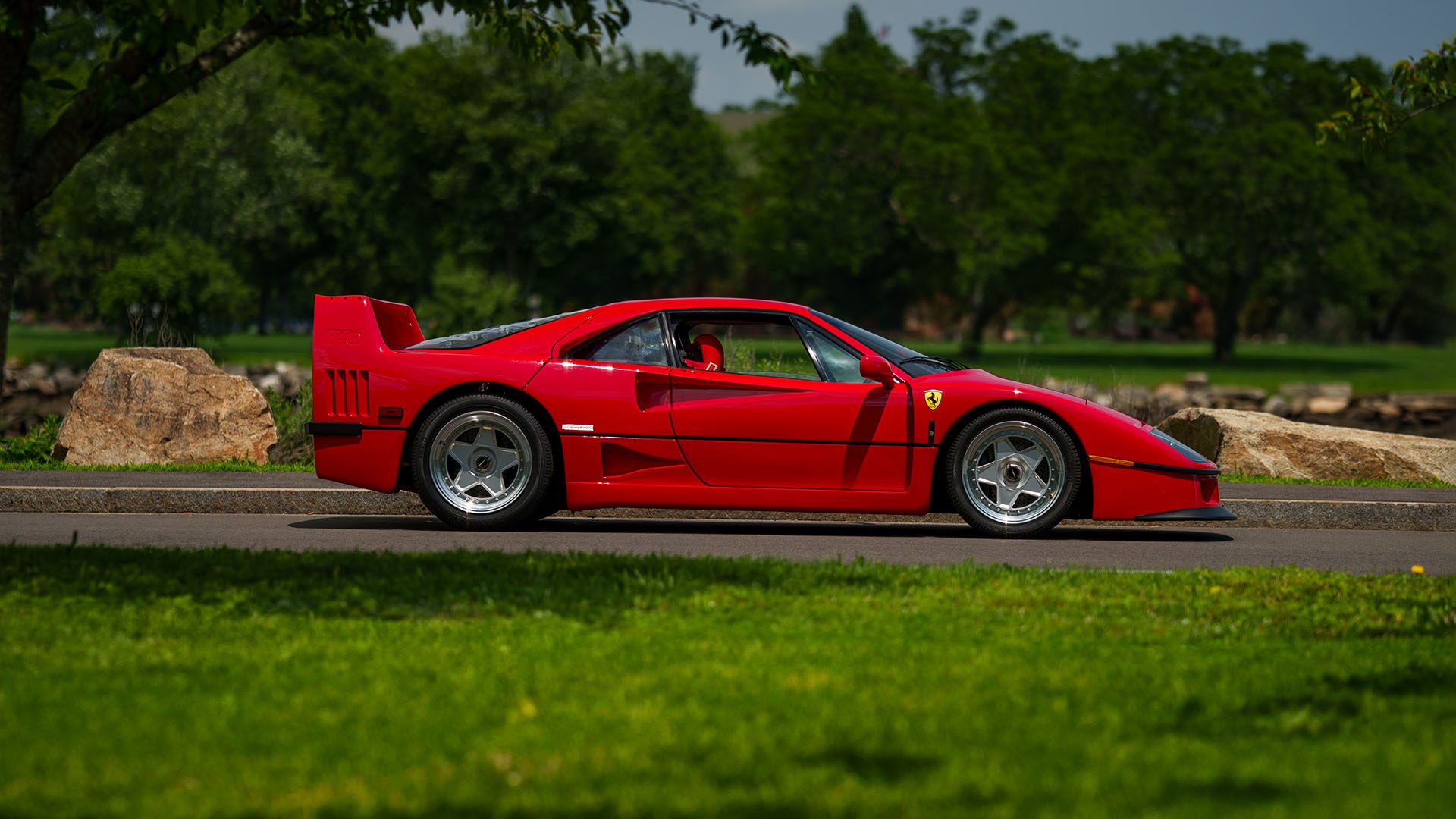 A red Ferrari F40 sports car is parked on a paved road, with grass, trees, and rocks in the background—an icon that could turn heads at the 2025 Monterey Auction by Broad Arrow.
