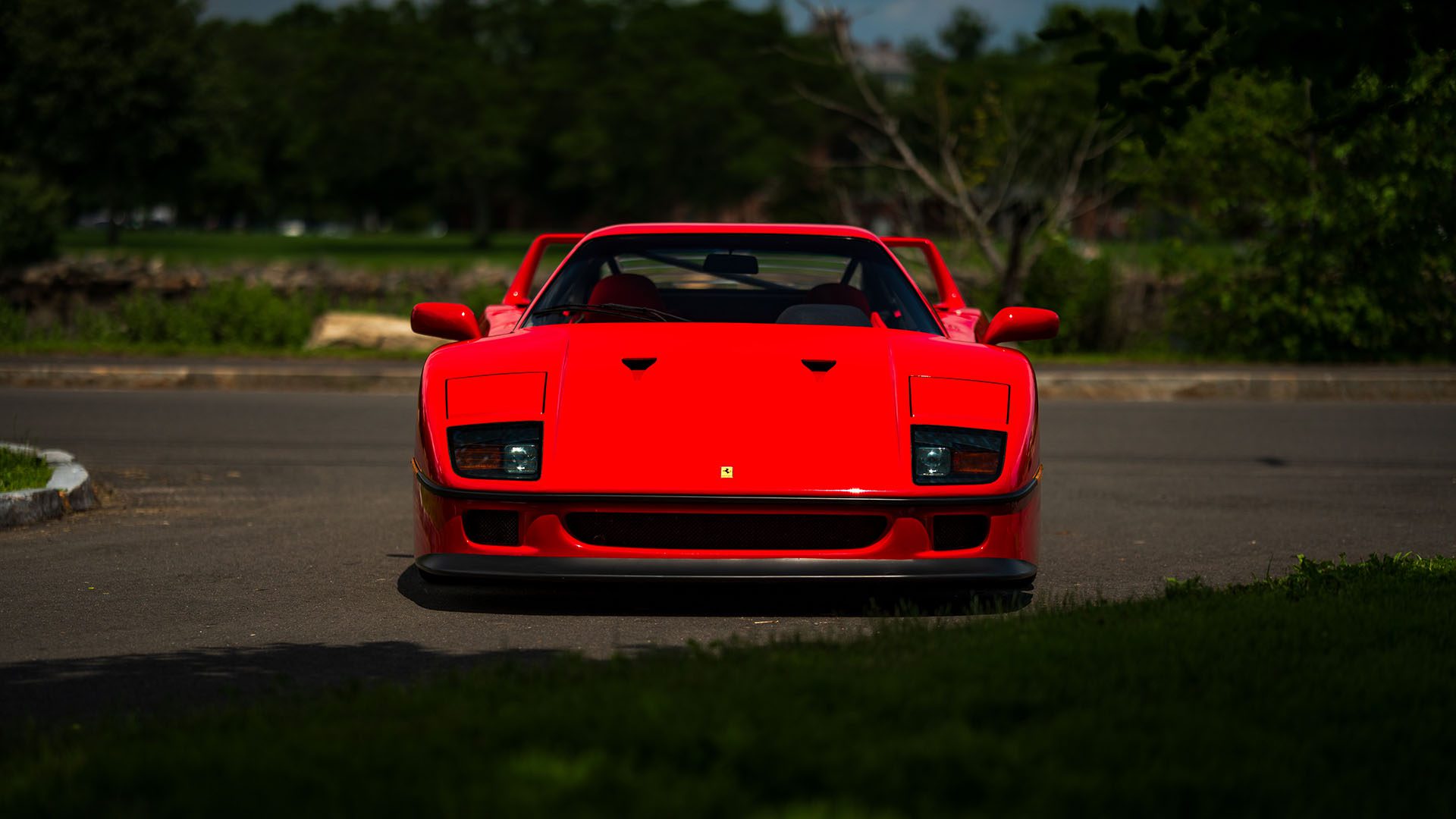 A red Ferrari F40, seen from the front, is parked on a paved road with green trees and grass in the background—a striking scene ahead of its appearance at the 2025 Monterey Auction by Broad Arrow.