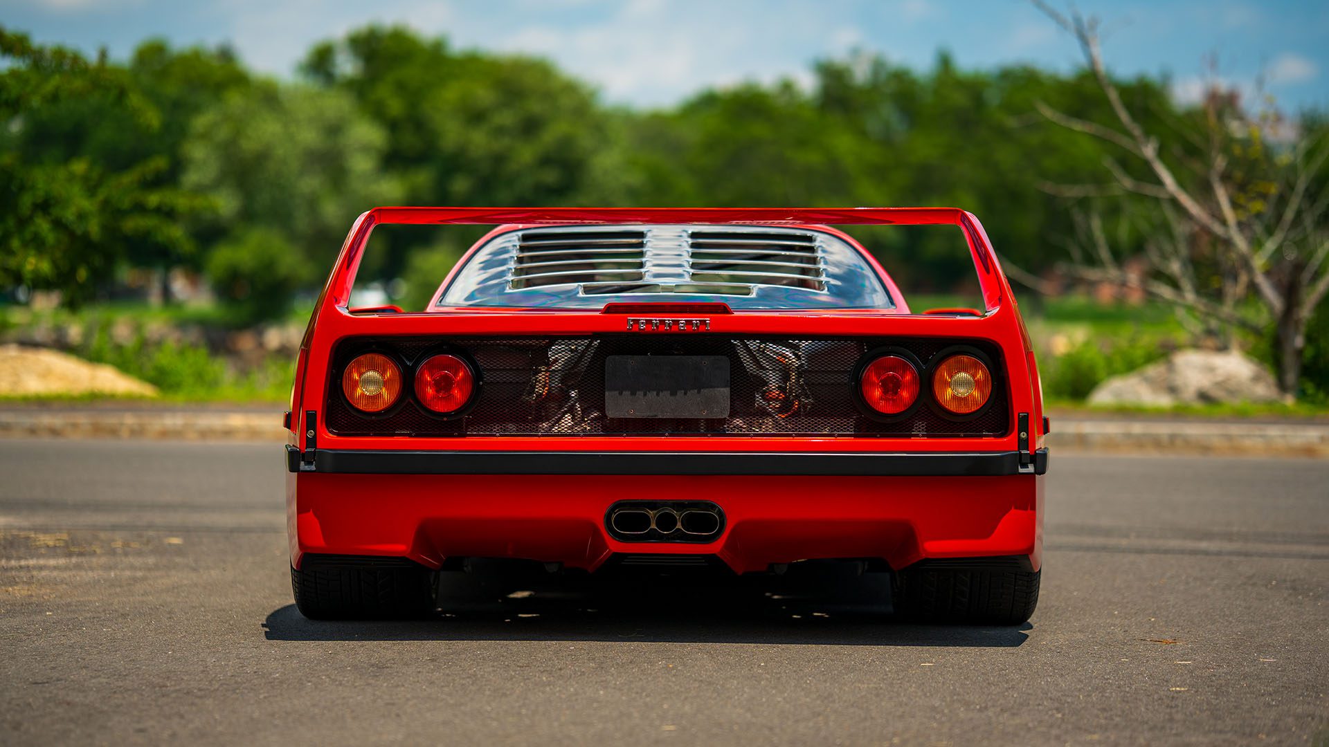 A red Ferrari F40 sports car viewed from the rear, parked on a paved surface with greenery and trees in the background under a sunny sky—an icon likely to draw attention at the 2025 Monterey Auction by Broad Arrow.