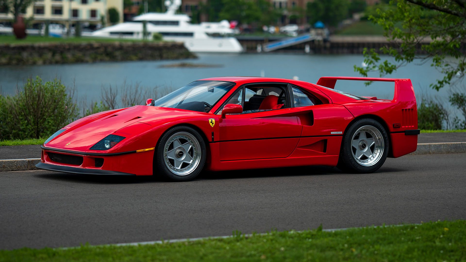 A red Ferrari F40 sports car is parked on the side of a road with water, a yacht, and buildings in the background, capturing the excitement of the upcoming Broad Arrow Monterey Auction.
