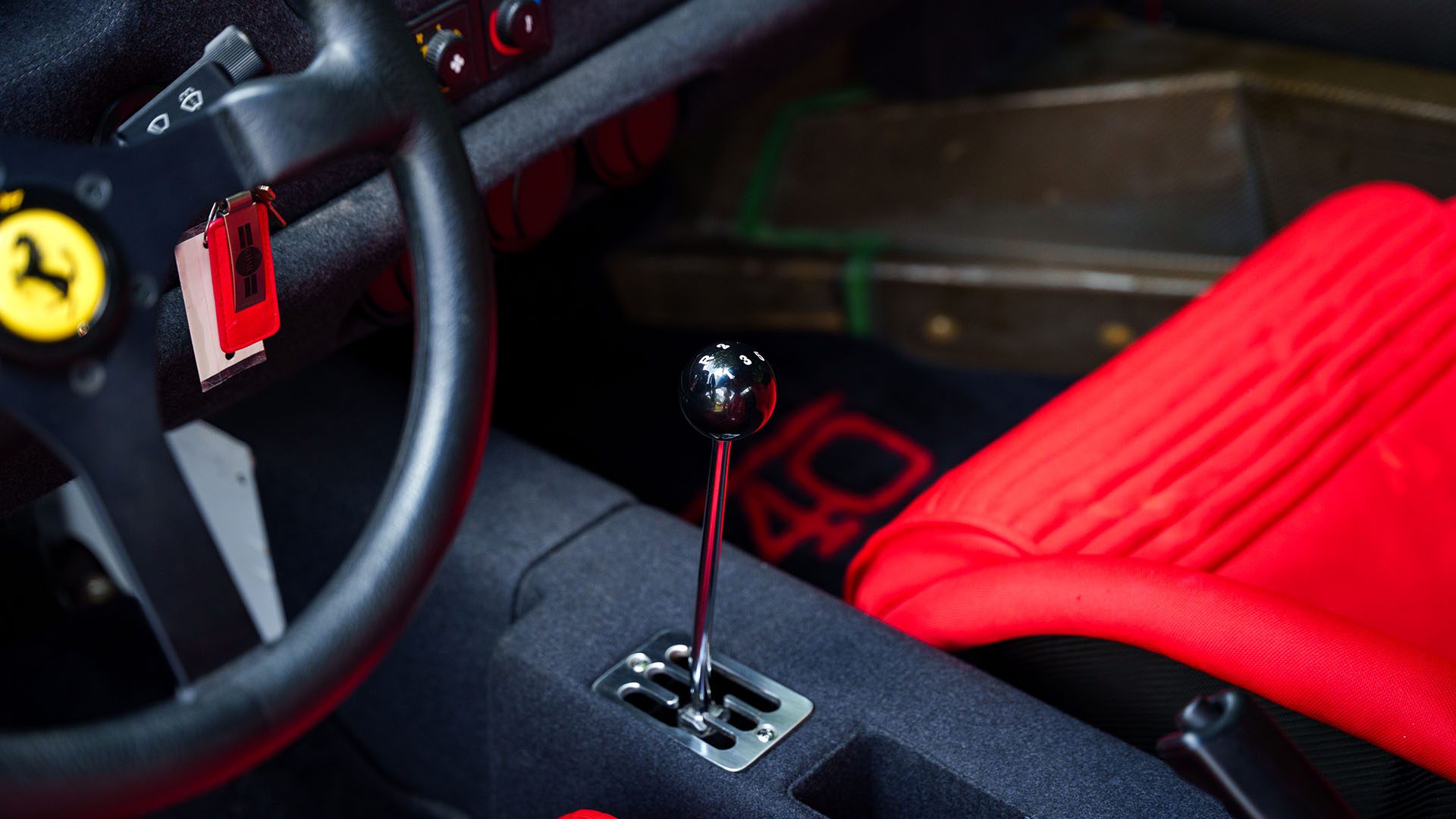 Close-up of a Ferrari F40 interior featuring a black manual gear shift, red seat, Ferrari logo on the steering wheel, and red key fob—captured ahead of the 2025 Monterey Auction by Broad Arrow.