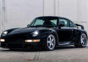 A black 1998 RUF CTR 2 Sport with a wide body kit and custom wheels is parked indoors against a white brick wall, ready to turn heads at the Monterey Jet Center Auction.