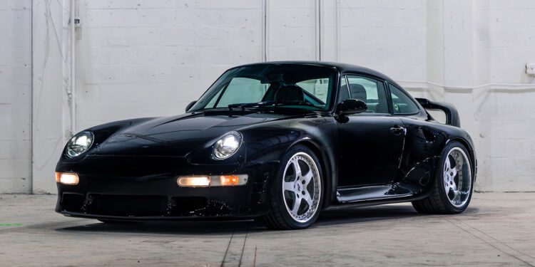 A black 1998 RUF CTR 2 Sport with a wide body kit and custom wheels is parked indoors against a white brick wall, ready to turn heads at the Monterey Jet Center Auction.
