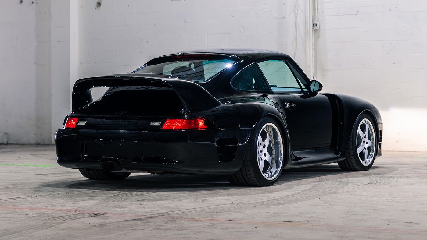 A black 1998 RUF CTR 2 Sport with a large rear spoiler is parked in a spacious, empty, white-walled garage.