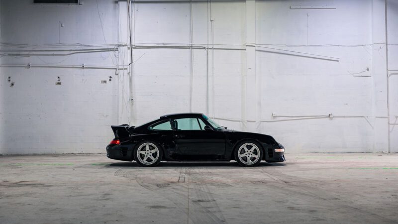 A black 1998 RUF CTR 2 Sport is parked in an empty, white-walled industrial space with concrete floors and exposed pipes, reminiscent of the Monterey Jet Center Auction setting.