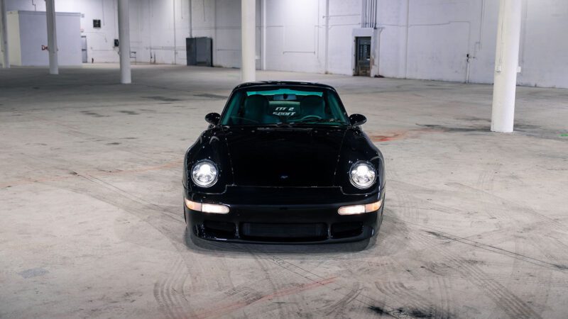 A 1998 RUF CTR 2 Sport black sports car is parked indoors in an empty, spacious warehouse with white walls and support columns, reminiscent of the Monterey Jet Center Auction setting.