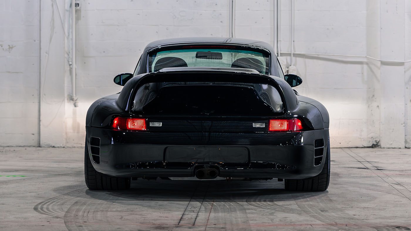 A black RUF CTR 2 Sport is parked indoors, viewed from the rear against a white wall with visible wiring and pipes—an impressive sight ahead of the Monterey Jet Center Auction by Broad Arrow.