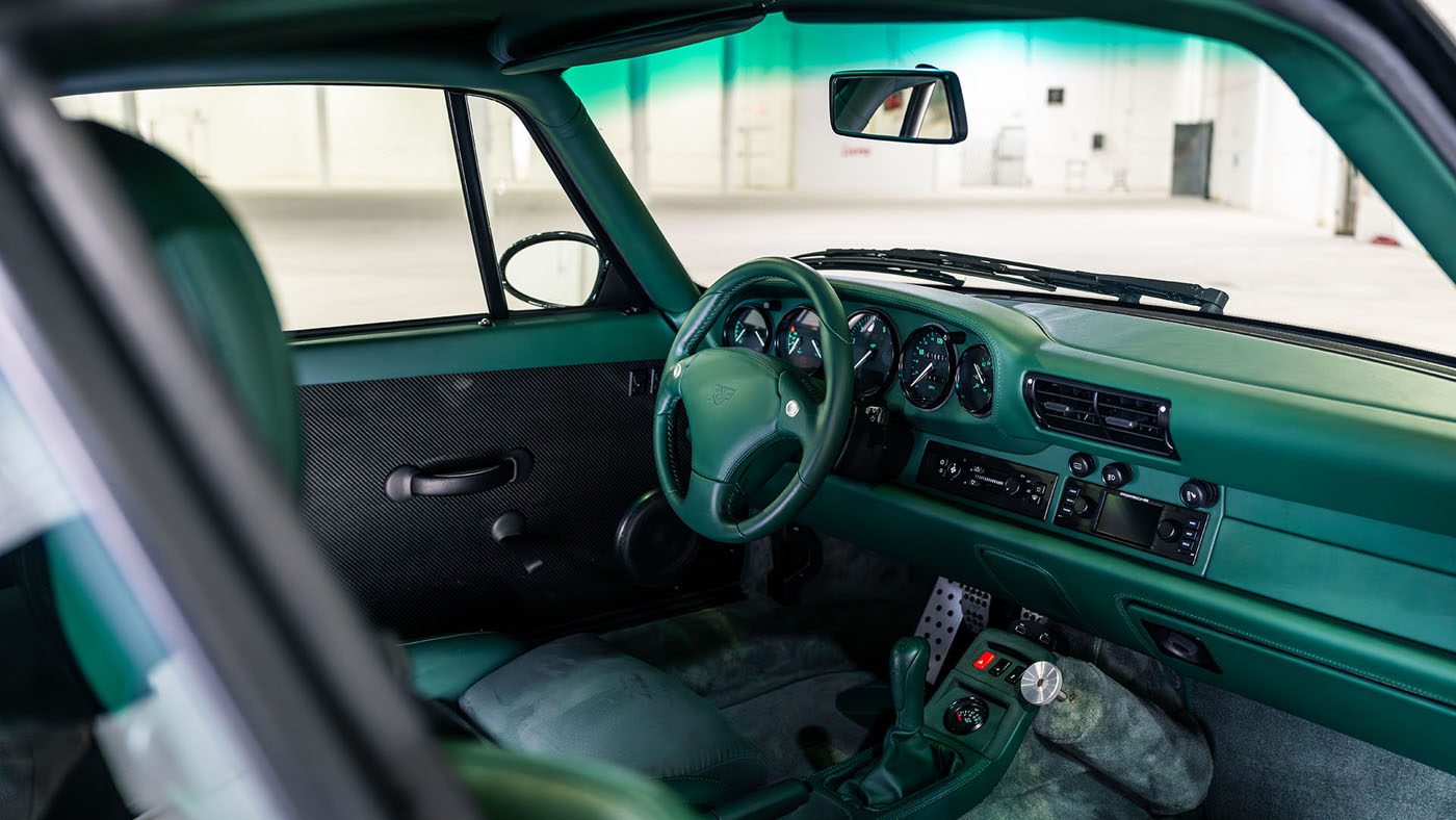 The image shows the green interior of a vintage car, focusing on the dashboard, steering wheel, manual gear shift, and front seats—possibly previewed at the Broad Arrow Monterey Jet Center Auction alongside gems like the RUF CTR 2 Sport.