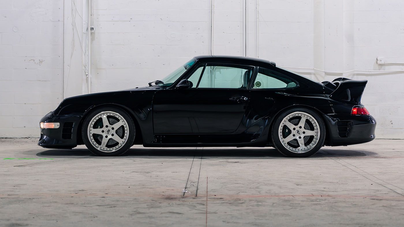 A black 1998 RUF CTR 2 Sport with a rear spoiler is parked indoors against a white brick wall, ready for the Monterey Jet Center Auction.