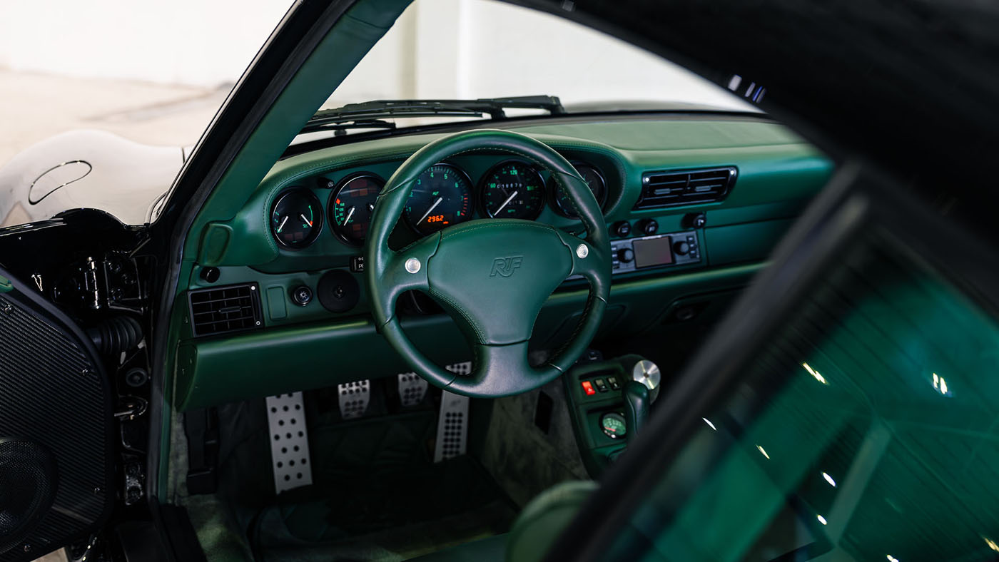 Interior view of a RUF CTR 2 Sport showing a green leather steering wheel with RUF logo, analog gauges, manual gear shifter, and metallic pedals—set to impress at the 2025 Monterey Jet Center Auction.