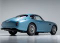 A vintage blue coupe, reminiscent of the DB4 GT Zagato Continuation, is parked in a brightly lit studio, shown from the rear left side with sleek lines and wire-spoke wheels.