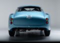 A light blue vintage sports car, reminiscent of classics seen at the Monterey Jet Center Auction, is shown from the rear against a plain white background, displaying its tail lights, dual exhaust pipes, and rear window.