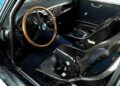 Interior of a classic sports car, like an Aston Martin DBS GT Zagato at the Monterey Jet Center Auction, featuring a wooden steering wheel, analog gauges, and two black carbon fiber racing seats with harnesses.