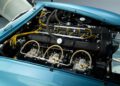 Close-up view of a classic car engine compartment with visible wiring, carburetors, and engine components under a blue hood, reminiscent of the DB4 GT Zagato Continuation showcased at the Monterey Jet Center Auction.
