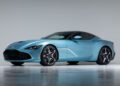 A light blue Aston Martin DBS GT Zagato two-door sports car with a black roof, silver wheels, and a sleek, aerodynamic design is parked in a studio with a plain gray background.