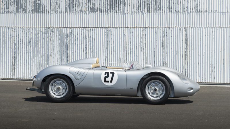 A vintage silver Porsche 718 RSK Spyder with the number 27 on the door is parked in front of a corrugated metal wall, recalling its glory days at the Monterey Jet Center Auction.