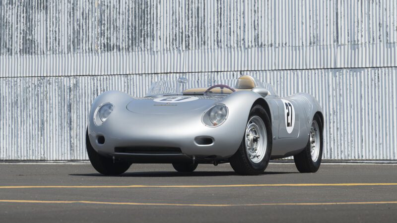 A silver vintage Porsche 718 RSK Spyder race car with the number 27 on the side is parked on an asphalt surface in front of a corrugated metal wall at the Monterey Jet Center Auction.