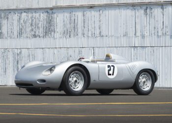 A silver Porsche 718 RSK Spyder vintage race car with the number 27 on its side is parked on an asphalt surface in front of a corrugated metal wall at the Monterey Jet Center Auction 2025.