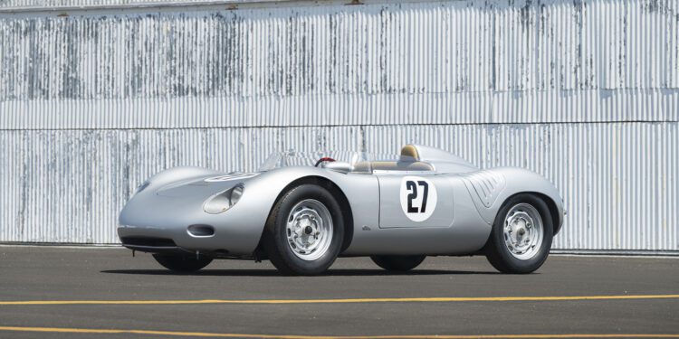 A silver Porsche 718 RSK Spyder vintage race car with the number 27 on its side is parked on an asphalt surface in front of a corrugated metal wall at the Monterey Jet Center Auction 2025.