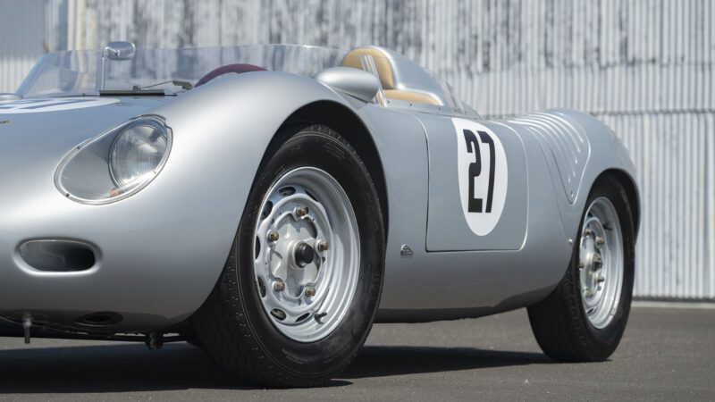 A silver Porsche 718 RSK Spyder vintage race car with the number 27 on the door is parked on pavement near a corrugated metal wall, ready for its spotlight at the Monterey Jet Center Auction.