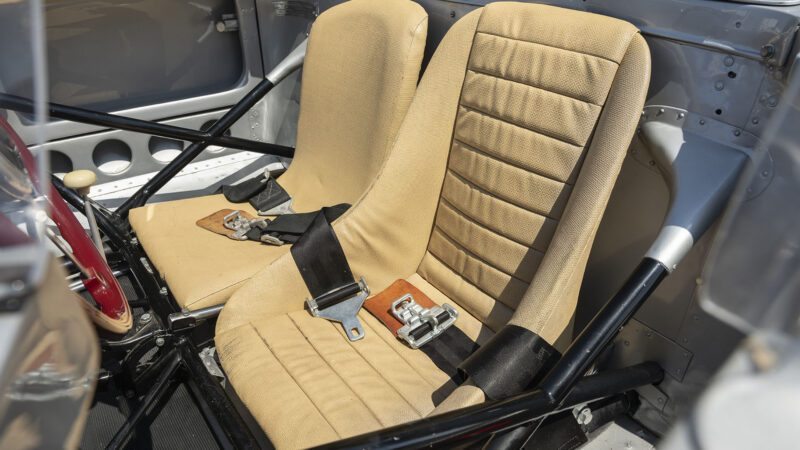 Two tan bucket seats with horizontal stitching and black seat belts are installed in a metal Porsche 718 RSK Spyder interior, featuring visible frame bars and a red steering wheel to the left. Monterey Jet Center Auction, 2025.