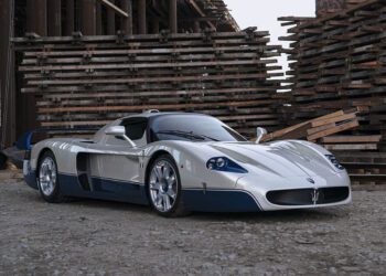 A striking white and blue 2005 Maserati MC12 sports car is parked on a gravel surface between large stacks of wooden planks and industrial beams at the Monterey Jet Center Auction.