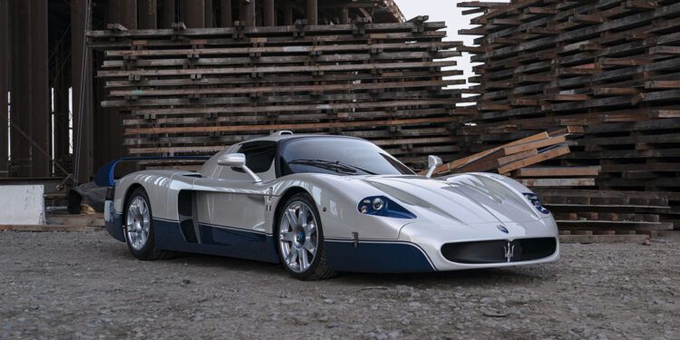 A striking white and blue 2005 Maserati MC12 sports car is parked on a gravel surface between large stacks of wooden planks and industrial beams at the Monterey Jet Center Auction.