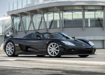 A black Koenigsegg CCXR sports car is parked on an empty road in front of a modern building with large glass windows, ready to turn heads at the 2025 Monterey Auction by Broad Arrow.