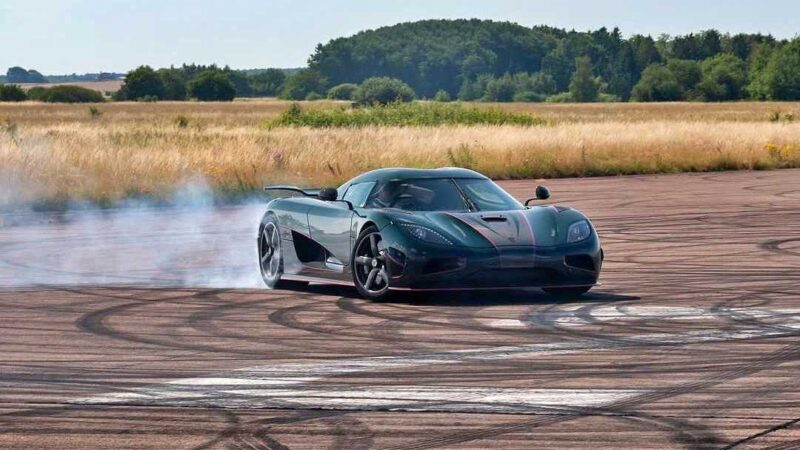 A sports car, reminiscent of the 1-of-1 Agera R reimagined by The Legends Division that Christian von Koenigsegg reveals, drifts on open asphalt, producing tire smoke with grassy fields and trees in the background.