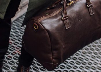 Person carrying a dark brown leather duffel bag with gold hardware, showcasing luxury accessories and effortless style while walking on a metal grate.