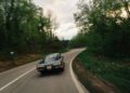 A vintage Lamborghini cruises along a winding road in Emilia-Romagna, surrounded by dense green trees under a cloudy sky.