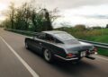 A vintage dark gray sports car, reminiscent of classic vintage Lamborghinis, is driving on a winding rural road bordered by trees and grass under a cloudy sky.