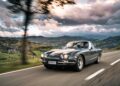 A classic black coupe, reminiscent of vintage Lamborghinis, drives along a scenic country road in Emilia-Romagna, with rolling hills and cloudy skies in the background.