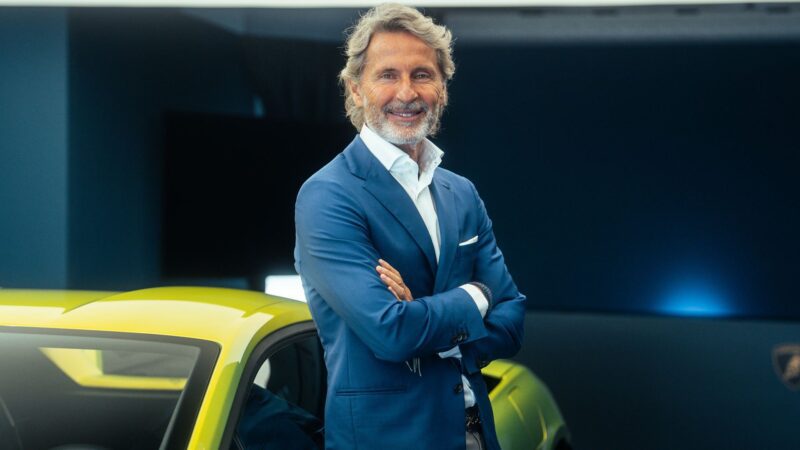 A man in a blue suit stands with arms crossed and smiles beside a yellow Lamborghini, celebrating record H1 2025 sales figures credited to the brand’s hybrid shift in a modern showroom.